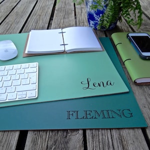 Może przedstawiać: Zielona sk&oacute;rzana podkładka na biurko z wygrawerowanym imieniem "Lena Fleming" czarnymi literami. Podkładka leży na drewnianej powierzchni z klawiaturą, notesem i telefonem kom&oacute;rkowym na g&oacute;rze.