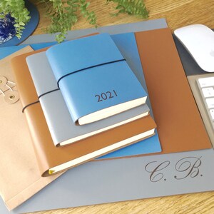 May include: A stack of four leather-bound journals in shades of brown and blue. The top journal is blue and has the year "2021" embossed on the cover. The journals are sitting on a gray desk mat with the initials "C.B." embossed in the bottom right corner.