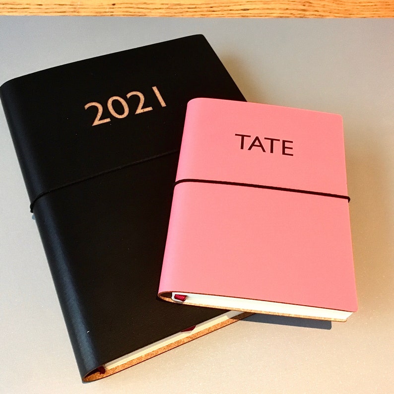 Pode incluir: Dois di&aacute;rios de couro preto, um com o ano 2021 gravado na capa e o outro com a palavra "TATE" gravada na capa. O di&aacute;rio com a palavra "TATE" &eacute; rosa.