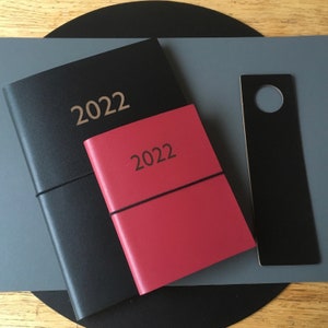 May include: Two black leather-bound journals with the year 2022 embossed on the front. The smaller journal is red and the larger journal is black. A black bookmark with a hole in the top is also visible.