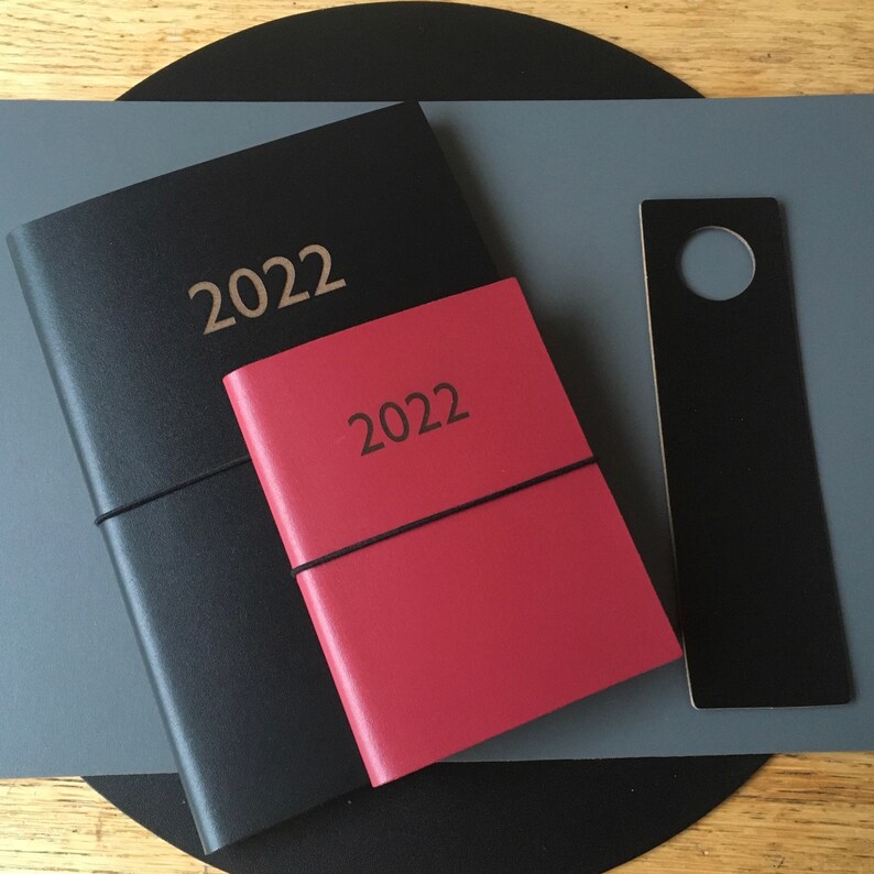 May include: Two black leather-bound journals with the year 2022 embossed on the front. The smaller journal is red and the larger journal is black. A black bookmark with a hole in the top is also visible.