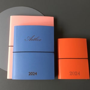 Pode incluir: Dois di&aacute;rios de capa dura azul e rosa com fechos el&aacute;sticos. O di&aacute;rio maior tem o texto "Artbox" e "2024" impresso na frente. O di&aacute;rio menor &eacute; laranja e tem o texto "2024" impresso na frente.