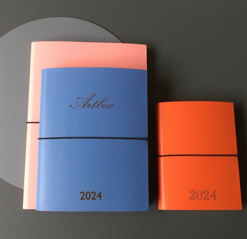 Pode incluir: Dois di&aacute;rios de capa dura azul e rosa com fechos el&aacute;sticos. O di&aacute;rio maior tem o texto "Artbox" e "2024" impresso na frente. O di&aacute;rio menor &eacute; laranja e tem o texto "2024" impresso na frente.