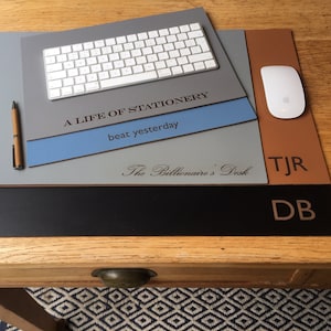 Może przedstawiać: Podkładka na biurko w kolorze szarym, niebieskim, brązowym i czarnym z tekstem "A Life of Stationery", "beat yesterday", "The Billionaire's Desk", "TJR" i "DB". Na podkładce znajduje się biała klawiatura i biała mysz.