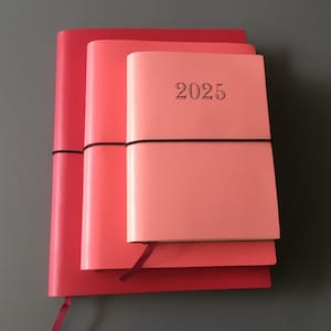 Pode incluir: Tr&ecirc;s di&aacute;rios de couro rosa e vermelho empilhados um em cima do outro. O di&aacute;rio de cima tem o ano "2025" impresso na capa.