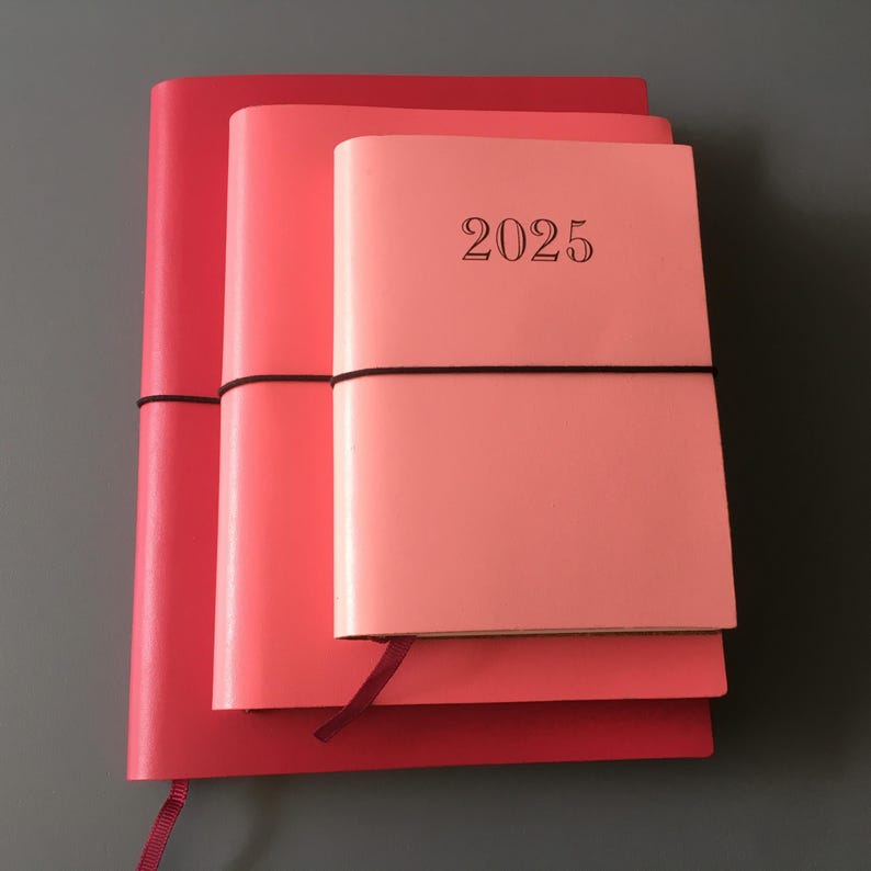Pode incluir: Tr&ecirc;s di&aacute;rios de couro rosa e vermelho empilhados um em cima do outro. O di&aacute;rio de cima tem o ano "2025" impresso na capa.