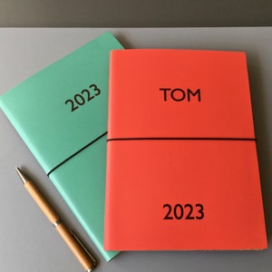 Pode incluir: Dois planejadores para 2023, um verde e um vermelho. O planejador vermelho tem o nome "TOM" impresso na capa.