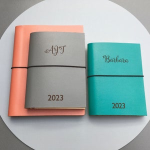 以下が含まれることがあります： ゴム留めの革装丁のジャーナルが2冊。グレーのジャーナルには「AJT」という名前と「2023」という年号が刻まれています。ターコイズブルーのジャーナルには「Barbara」という名前と「2023」という年号が刻まれています。