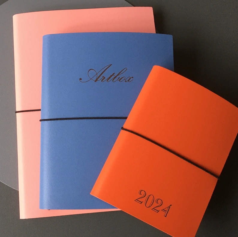 May include: Three notebooks stacked on top of each other. The top notebook is orange and has the year 2024 printed on it. The middle notebook is blue and has the word "Artbox" printed on it. The bottom notebook is pink.