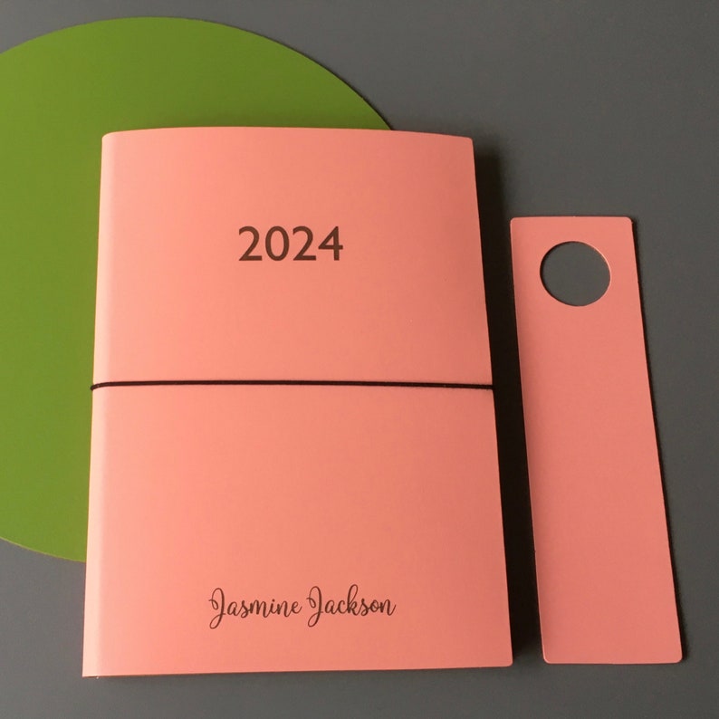 May include: A pink 2024 planner with a black elastic band and the name "Jasmine Jackson" printed on the front. The planner has a matching pink bookmark with a hole in the top.