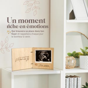 Op de afbeelding: Een houten fotolijst met een echografie, de tekst "TU VAS &Ecirc;TRE Mamie" en "Juin 2025". De lijst staat open op een witte plank, met Franse tekst op de muur erboven.