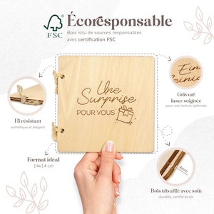 Op de afbeelding: Een houten vierkante geschenkdoos met de tekst "Une Surprise POUR VOUS" en een cadeau-illustratie. De doos is gemaakt van lichtgekleurd hout en wordt door een hand vastgehouden. De doos is 14x14 cm.