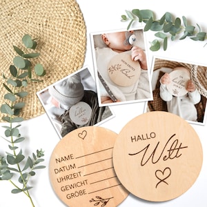 May include: Two wooden baby milestone discs. One disc has the text "HALLO Welt" with a heart design. The other disc has the text "NAME, DATUM, UHRZEIT, GEWICHT, GR&Ouml;BE" with a heart and a small branch design.