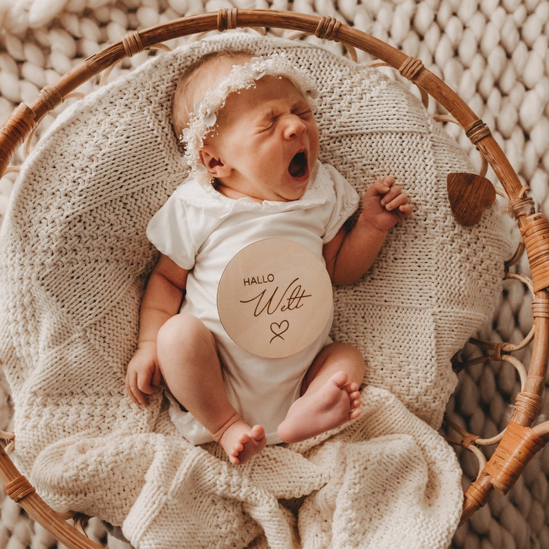 May include: A newborn baby wearing a white onesie with a wooden sign that says "Hallo Welt" with a heart symbol. The baby is lying in a wicker basket with a white blanket.