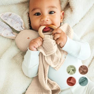 May include: A baby is holding a beige muslin bunny rattle with a wooden ring. The bunny has a small pink heart on its chest. The baby is wearing a light blue long-sleeved shirt.