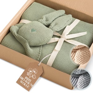 May include: A green knitted baby blanket, a matching knitted bunny toy, and a pair of knitted mittens are packaged in a brown box with a white ribbon. A tag attached to the box reads "100% BIO-BAUM WOLLE".