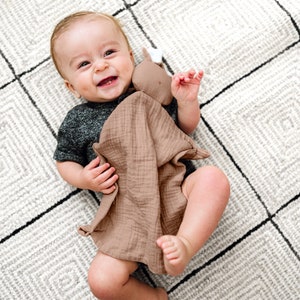 Peut inclure: Un bébé allongé sur un tapis à carreaux blancs et bruns, tenant un jouet en mousseline brun avec une petite tête d'animal.