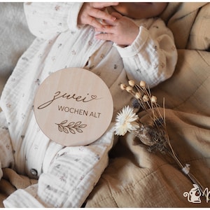 May include: A wooden baby milestone sign with the text "zwei Wochen alt" (two weeks old) in German, with a small heart and a leaf design. The sign is on a tan blanket with a baby wearing a white onesie.