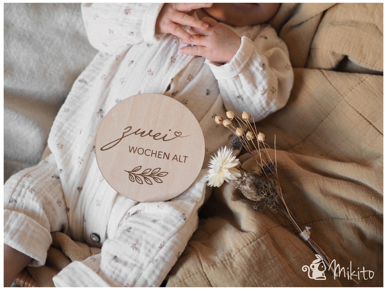 May include: A wooden baby milestone sign with the text "zwei Wochen alt" (two weeks old) in German, with a small heart and a leaf design. The sign is on a tan blanket with a baby wearing a white onesie.
