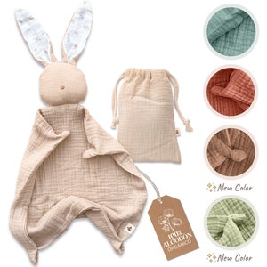 May include: A beige organic cotton muslin bunny lovey with a white floral print on the ears. The lovey comes with a matching beige muslin storage bag and a tag that reads "100% ALGODON ORGANICO".
