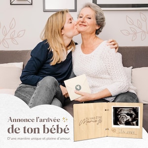 Op de afbeelding: Houten aankondiging met de tekst "Tu vas &ecirc;tre Mamie" en een echo-afbeelding van juni 2025. Twee vrouwen omarmen elkaar. De tekst "Annonce l'arriv&eacute;e de ton b&eacute;b&eacute;" is ook zichtbaar.
