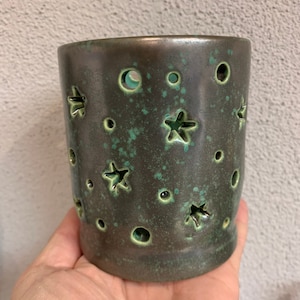 Può includere: Un portacandele in ceramica verde scuro con ritagli a forma di stelle e cerchi. Il portacandele ha una finitura maculata e un interno turchese. La forma cilindrica è tenuta in una mano.