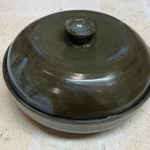 Può includere: Un piatto rotondo in ceramica con coperchio, di colore verde oliva scuro con una finitura liscia e lucida. Il coperchio presenta un piccolo pomello centrale. La ciotola ha una base di un verde più chiaro e spento, di forma circolare.
