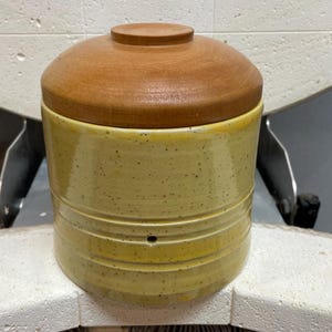 Könnte beinhalten: Eine gelbe Keramikdose mit einem Holzdeckel. Die Dose hat eine gesprenkelte Glasur und drei horizontale Linien um die Mitte. Der Deckel ist hellbraun.