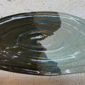 Könnte beinhalten: Eine ovale Keramikplatte mit glänzender Oberfläche und einer deutlichen Farbtrennung. Eine Hälfte ist in einem tiefen, dunklen Anthrazitbraun gehalten, während die andere Hälfte in einem hellen, gesprenkelten Salbeigrün erscheint. Die Oberfläche weist konzentrische Rillen auf.