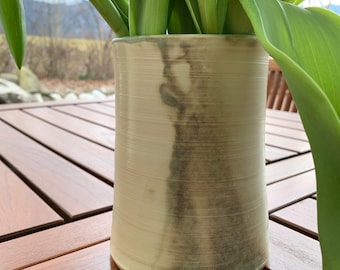 Può includere: Un vaso in ceramica beige chiaro con una finitura testurizzata simile al legno. Il vaso ha una leggera glassa verde sul fondo ed è appoggiato su una superficie in legno.