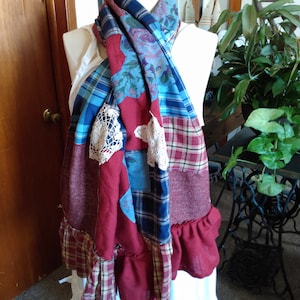 May include: A patchwork scarf with a variety of fabrics, including plaid, floral, and solid burgundy. The scarf has a ruffled edge and lace accents, displayed on a mannequin. The scarf is a mix of blues, reds, and whites.