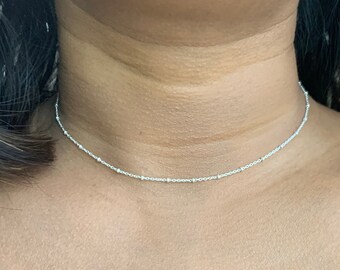 May include: A delicate silver chain choker necklace with small beads. The necklace is worn by a person with a red shirt.