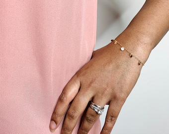 May include: A gold-colored bracelet with small, round charms is worn on a person's wrist. The bracelet is delicate and simple, with several charms evenly spaced along the chain. The person is wearing a pink top and has multiple rings on their finger.