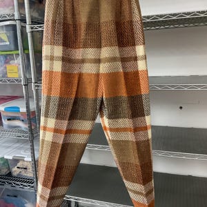 May include: Brown, orange, and cream plaid pants hanging on a white hanger. The pants have a high waist and a straight leg. The plaid pattern is made up of horizontal and vertical lines, creating a classic look.