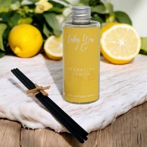 May include: A clear glass bottle with a silver cap and a yellow label that reads "Lakes Way & Co Sparkling Lemon Reed Diffuser Refill." Black diffuser sticks are tied with a small brown bow. Lemons are in the background.