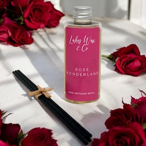 May include: A clear glass bottle with a silver cap and a pink label that reads "Lakes Wax & Co ROSE WONDERLAND REED DIFFUSER REFILL." Black reed sticks are tied with a tan ribbon. Red roses are scattered around the bottle.