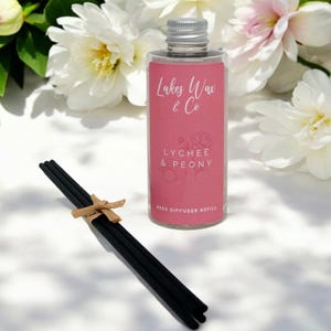 May include: A clear glass reed diffuser refill bottle with a silver lid and a pink label. The label reads "Lakes Wax & Co" and "Lychee & Peony". A bundle of black reeds tied with twine sits in front of the bottle. White and pink flowers are in the background.