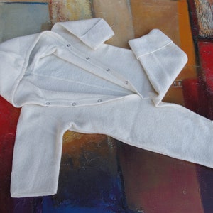 May include: A white hooded baby onesie with a zipper closure and button snaps. The onesie is made of a soft, fuzzy material and is laid out on a colorful abstract background.