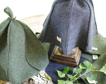 May include: Two felted wool cloche style hat covers in green and gray. The hats are sitting on a wooden table with a piece of birch bark and a sprig of birch leaves.