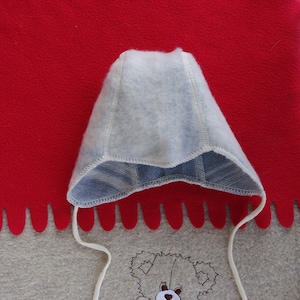 May include: A white baby bonnet with blue trim and white ties. The bonnet is lying on a beige fabric with a brown embroidered teddy bear design.