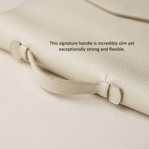 May include: Close-up of a cream-colored leather bag with a slim handle. The handle is attached with small, matching leather loops and stitching. The bag's surface has a textured appearance. Text on the image reads: "This signature handle is incredibly slim yet exceptionally strong and flexible."