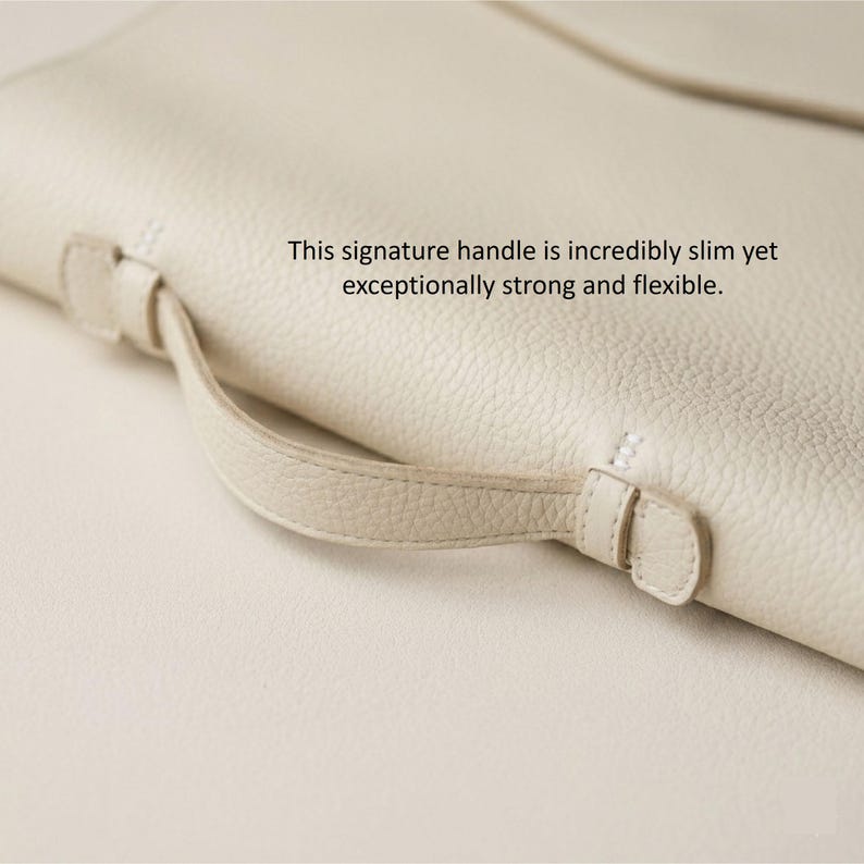 May include: Close-up of a cream-colored leather bag with a slim handle. The handle is attached with small, matching leather loops and stitching. The bag's surface has a textured appearance. Text on the image reads: "This signature handle is incredibly slim yet exceptionally strong and flexible."