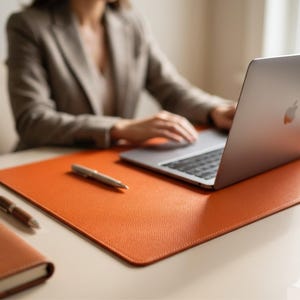 Pode incluir: Um tapete de secretária de couro laranja com um caderno e uma caneta a condizer. Uma caneta prateada repousa sobre o tapete perto de um computador portátil. Os acessórios de secretária estão numa secretária de cor clara.