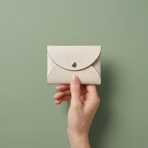 May include: A small, rectangular, off-white card holder with a textured surface and a rounded flap closure. The accessory features a single silver-toned metal snap button centered on the flap, providing a secure fastening for the folded design.