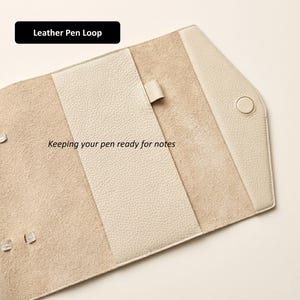 May include: Cream-colored leather pen loop with a snap closure. The interior features a soft, suede-like texture, with a strap to hold a pen. The text "Leather Pen Loop" and "Keeping your pen ready for notes" are visible.