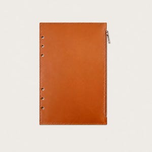 Può includere: Un inserto rettangolare per agenda in pelle color cuoio, dotato di una cerniera argentata verticale sul bordo destro e di una serie di sei fori circolari punzonati sul lato sinistro per l'aggancio, con cuciture visibili lungo il perimetro.