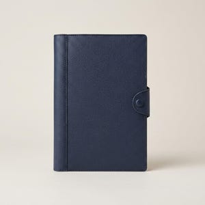 May include: A navy blue leather journal with a textured surface. The journal has a button closure on the right side and a vertical seam on the left side. The cover is a solid, dark blue color.