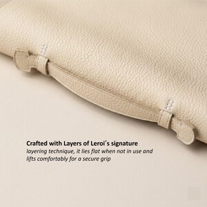 May include: Close-up of a cream-colored leather item with a handle. The handle is attached with small straps and stitching. The text in the image reads: Crafted with Layers of Leroi's signature layering technique, it lies flat when not in use and lifts comfortably for a secure grip.