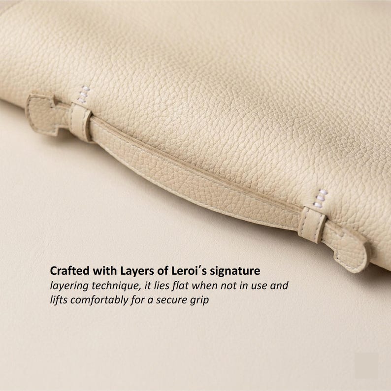 May include: Close-up of a cream-colored leather item with a handle. The handle is attached with small straps and stitching. The text in the image reads: Crafted with Layers of Leroi's signature layering technique, it lies flat when not in use and lifts comfortably for a secure grip.