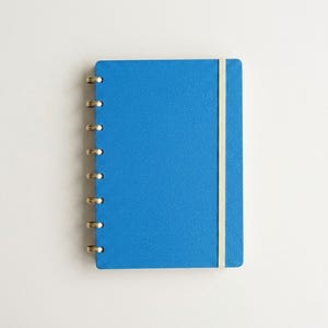 Könnte beinhalten: Ein leuchtend blaues Notizbuch mit texturiertem Einband und einem vertikalen Gummibandverschluss. Das Notizbuch verfügt über eine goldfarbene Metall-Discbinding-Bindung an der linken Seite, die das Hinzufügen oder Entfernen von Seiten ermöglicht.